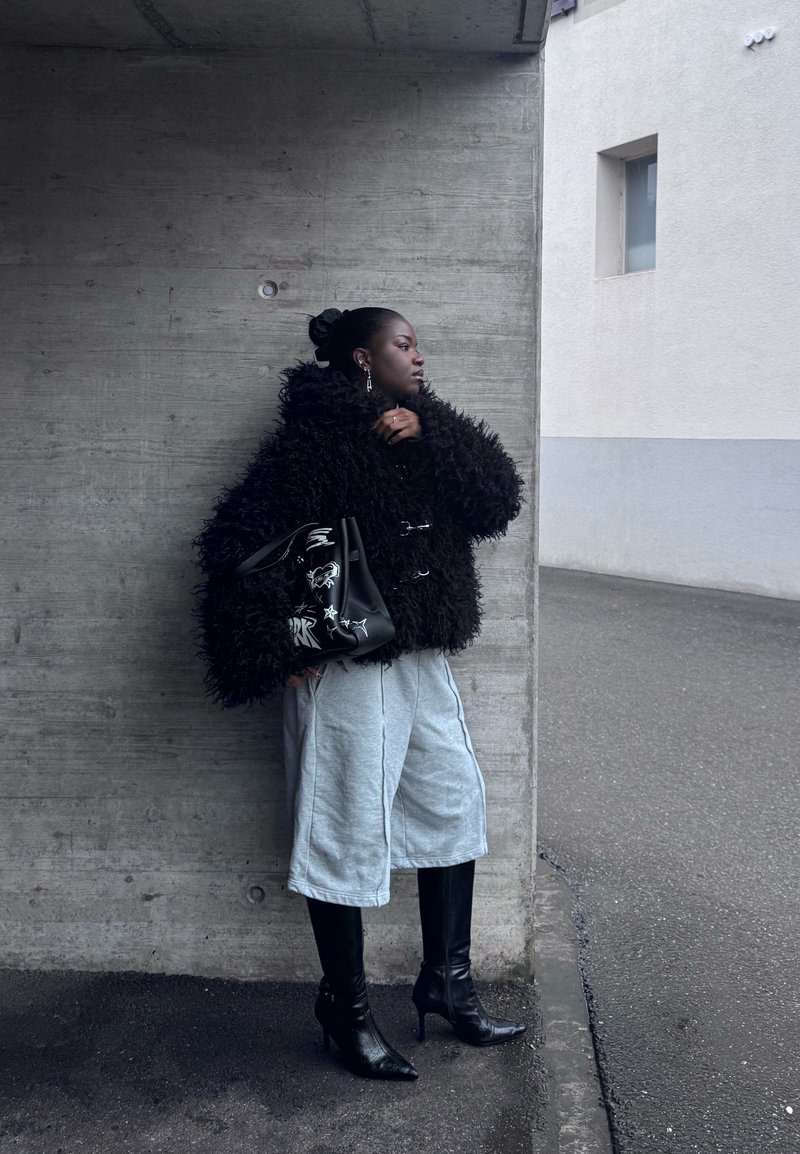 Femme en veste noire en matière duveteuse, short large gris et bottes à talons noires, debout contre un mur en béton, tenant un sac à main noir avec des motifs blancs.