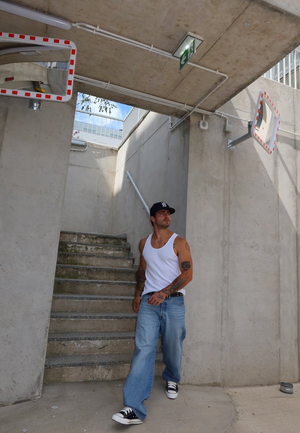 Hombre con tatuajes que lleva una camiseta sin mangas blanca y vaqueros azules bajando unas escaleras de cemento en un entorno urbano con dos espejos convexos y un cartel de salida arriba.