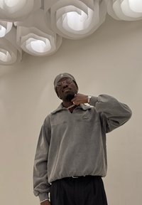 Grauer Pullover mit Kragen, versehen mit einer Naht auf der Brust und Logodetail, kombiniert mit dunkel gestreifter Hose, vor neutralem Hintergrund.