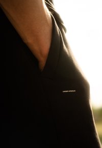 Čierny športový dres s dizajnom do V, so malým bielym logom "UNDER ARMOUR" na boku. Hladká textúra, detail na vrecku.