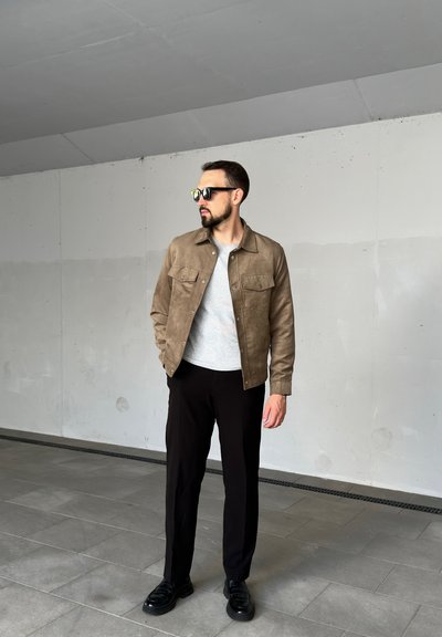 Chaqueta de ante marrón con bolsillos frontales, suéter gris, pantalones negros y mocasines negros, sobre un fondo de concreto liso.