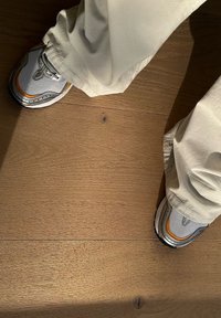 Graue und orange Sportchuhe mit einem oberen Teil aus Netzmaterial und silbernen Akzenten, kombiniert mit hellcremefarbenen Hosen, die auf einem Holzboden liegen.