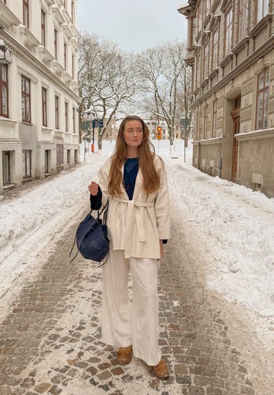 Mujer joven con abrigo y pantalones beige de pie en una calle adoquinada nevada entre edificios altos, sosteniendo una bolsa azul oscuro.