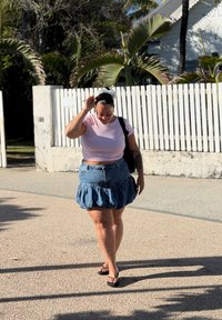 Lyserød tætsiddende T-shirt, denimnederdel med flæser, sorte flip-flops og et hvidt pandebånd. Udendørs omgivelser med palmer og et hvidt hegn.