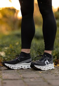 Inimene, kes kannab musti retuuse ja musti jooksujalatseid valgete paksude talladega, seisab väljas sillutatud rajal.