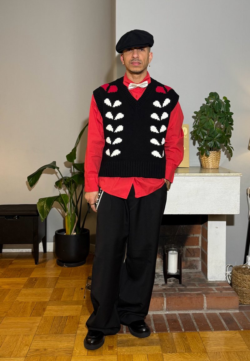 Personne portant un béret noir, un gilet à motifs noirs et blancs sur une chemise rouge avec un nœud papillon blanc, pantalon noir, debout à l'intérieur près de plantes.