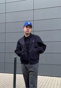 Tengerészeti bomber dzseki fekete pólóval, szürke nadrággal és kék LA baseball sapkával, áll egy szürke fal előtt.