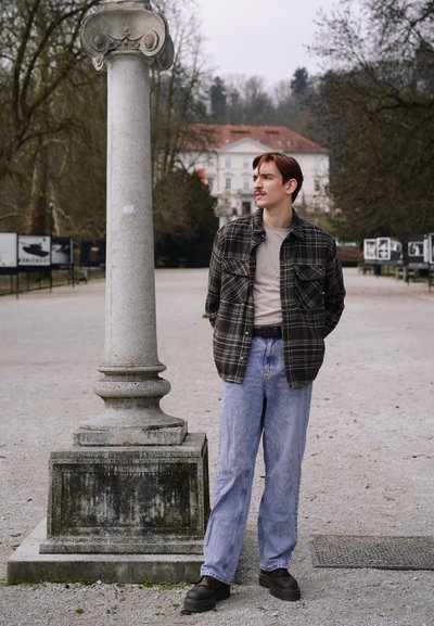 Joven con ropa informal de pie con las manos detrás de la espalda cerca de una columna de piedra en un parque al aire libre con árboles y un edificio de fondo.