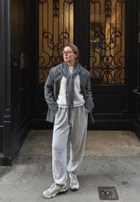 Blazer en laine grise avec une écharpe grise douce, un pull blanc et un pantalon de survêtement gris. Les baskets présentent un mélange de gris et d'accents clairs. Fond de porte décorative.