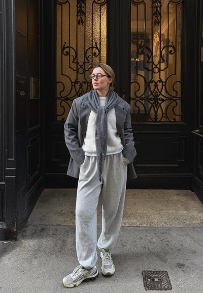 Blazer de lana gris con una bufanda gris suave, suéter blanco y pantalones deportivos grises. Las zapatillas tienen una mezcla de tonos grises y acentos claros. Fondo con una puerta decorativa.