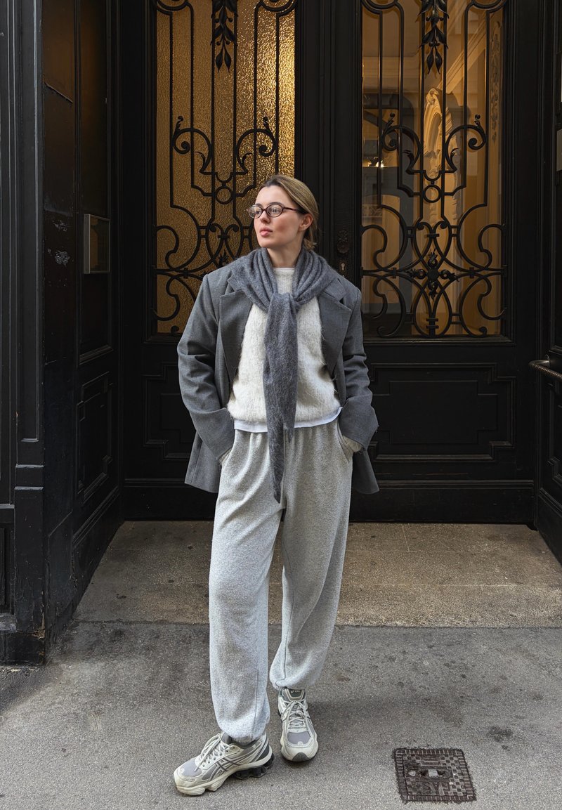 Blazer en laine grise avec une écharpe grise douce, un pull blanc et un pantalon de survêtement gris. Les baskets présentent un mélange de gris et de touches claires. Fond de porte décorative.