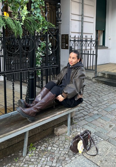 Mujer joven con chaqueta de cuero marrón y botas sentada en un banco junto a una elaborada puerta de hierro negra con un bolso marrón sobre el suelo empedrado.