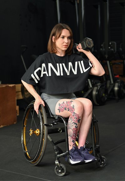 Mujer en silla de ruedas levantando una pesa con un brazo, vistiendo una camiseta negra con la inscripción "ICANIWILL" y mostrando un tatuaje floral en la pierna en un entorno de gimnasio.