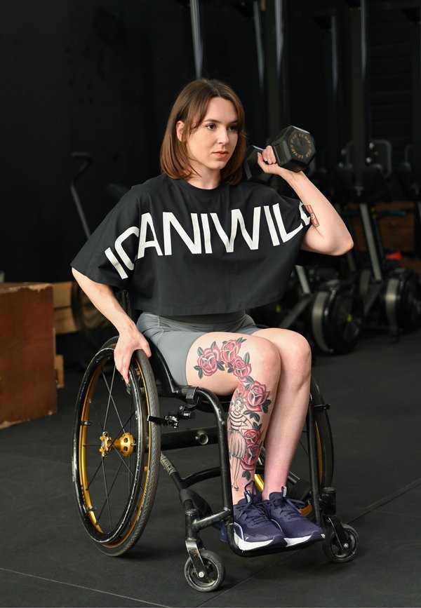 Kvinde i kørestol løfter en håndvægt med den ene arm, iført en sort "ICANIWILL" t-shirt og viser en blomster-tatovering på benet i et fitnessmiljø.