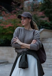 Rjav pleten pulover, bela satenska krila, zelena karirana srajca zavezana okoli pasu, rjav klobuk, rjava torba za prenos. Ozadje ima zamegljeno zelenje.