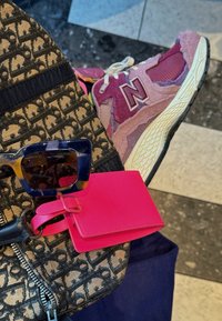 Rožinės New Balance sportbačiai su tekstūruotais akcentais, raštuota juoda audinio pirkinių maišelis, tamsiai mėlynas fonas, dideli kvadratiniai mėlyni akiniai nuo saulės ir rožinis bagažo žymeklis.