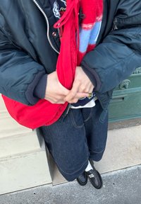 Personne portant une veste noire et un jean large tenant un sac rouge, debout près d'une porte verte les mains jointes devant.