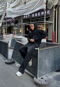 Sudadera negra con cremallera, pantalones anchos negros con rayas verticales, zapatillas grises, sentado en una barrera de metal con superficie texturizada, fondo urbano.