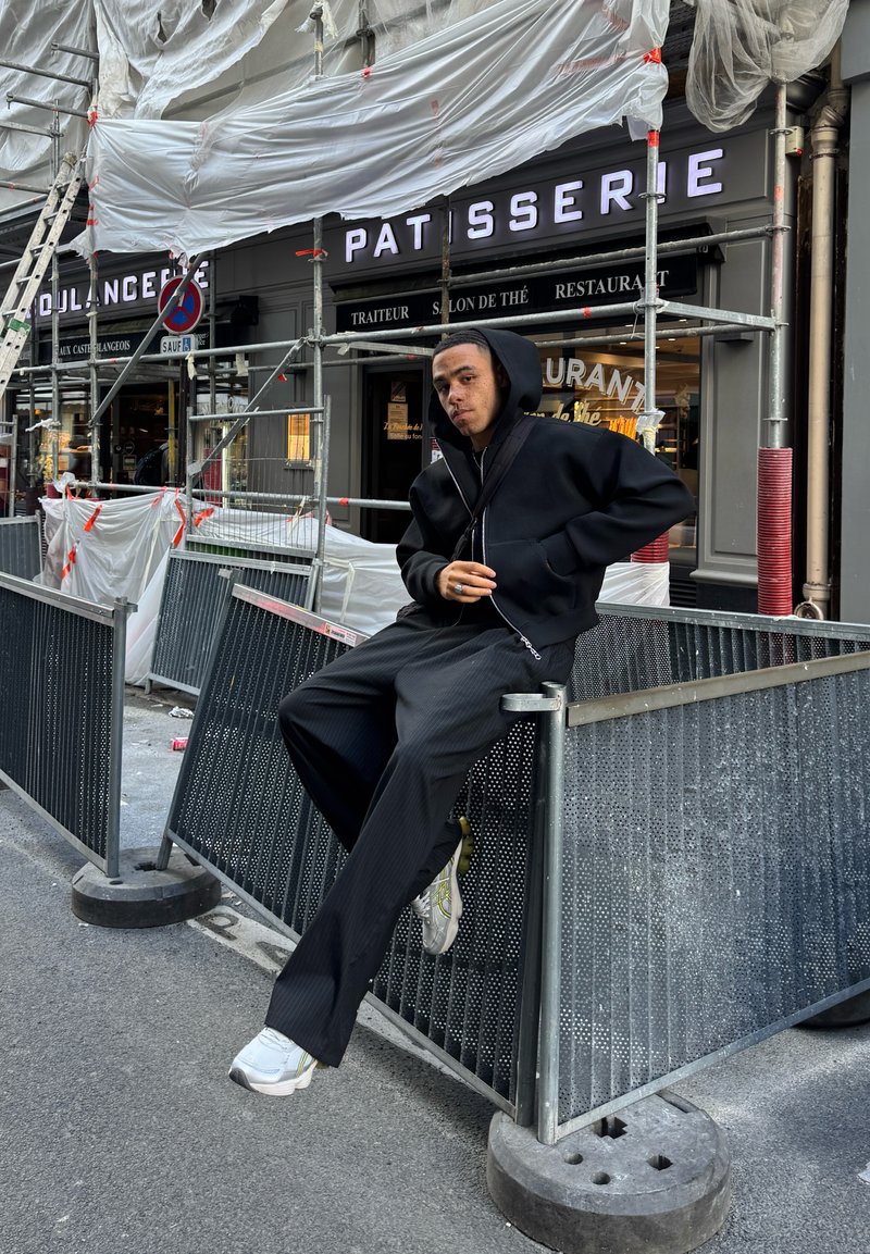 Black zip-up hoodie, wide black pants with vertical stripes, gray sneakers, seated on a metal barrier with textured surface, city background.