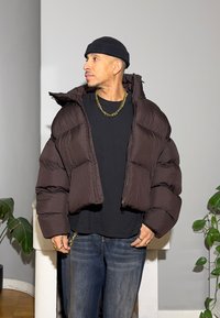 Ruskea toppatakkimallinen takki, jonka leikkaus on lyhyt ja jossa on suuret tikatut osiot sekä korkea kaulus, yhdistettynä mustaan paitaan ja sinisiin farkkuihin.