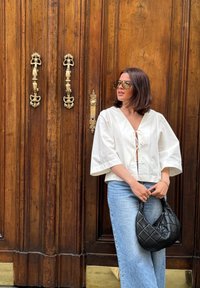 Άσπρη, χαλαρή μπλούζα με V λαιμόκοψη και λεπτομέρεια με κορδόνι, συνδυασμένη με ανοιχτό μπλε φαρδιά jeans. Μαύρη επενδυμένη τσάντα κρατημένη στα χέρια.