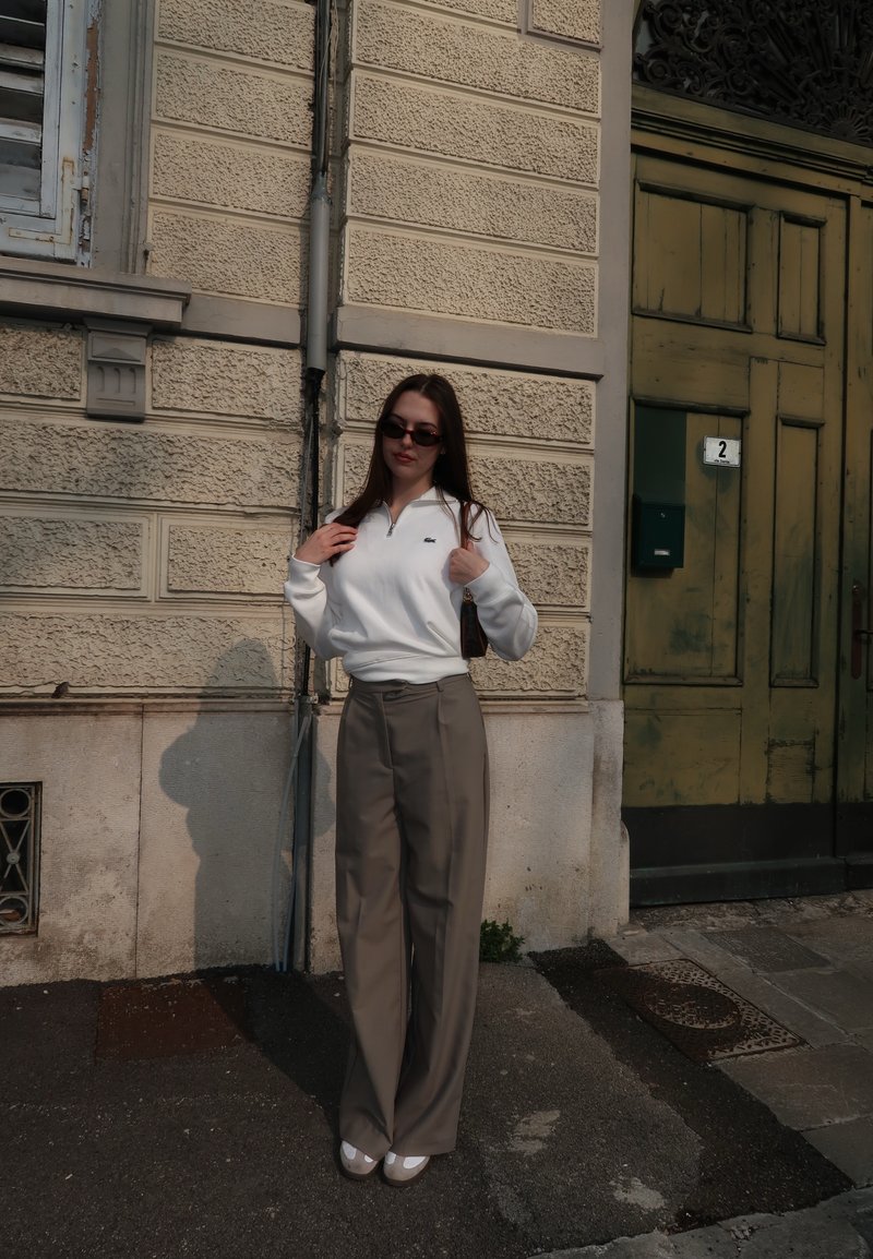 Femme portant des lunettes de soleil, un haut blanc à manches longues, un pantalon beige à jambes larges et des chaussures blanches, se tenant devant un mur beige texturé et une porte verte.