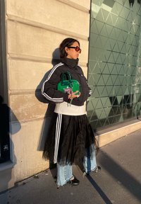 Femme portant une doudoune noire à rayures, jupe en tulle noire par-dessus un jean, lunettes de soleil orange, tenant un sac à main vert, debout près d'un mur beige et d'une vitre.