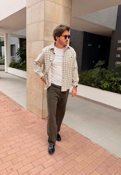Hombre con gafas de sol, camisa blanca, chaqueta de cuadros beige, pantalones y zapatos negros caminando por un pavimento de ladrillo cerca de una columna con azulejos y plantas.