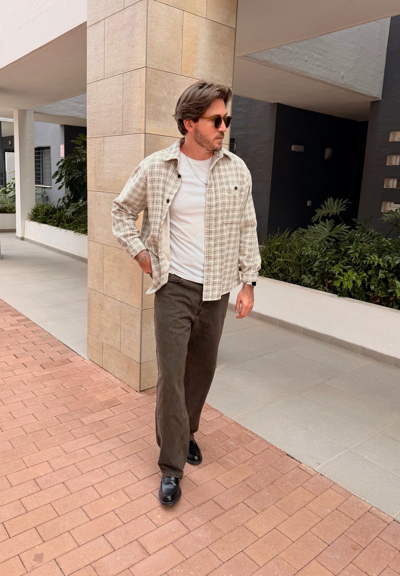 Homme portant des lunettes de soleil, une chemise blanche, une veste à carreaux beige, un pantalon et des chaussures noirs, marchant sur un pavé en briques près d'une colonne carrelée et des plantes.