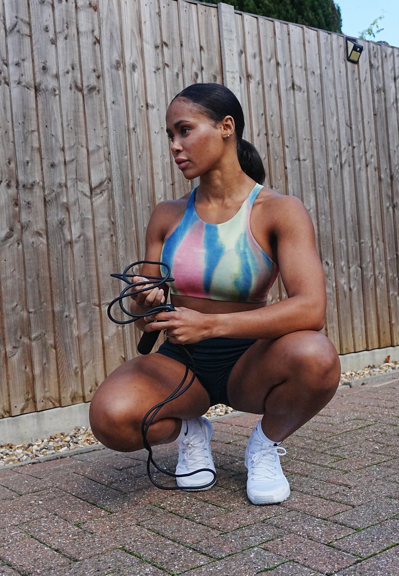 Top athletic scurt, cu un model tie-dye multicolor, pantaloni scurți negri, ținând o frânghie pentru sărit, ghemuit pe un paviment de cărămidă, cu un fundal de gard din lemn.