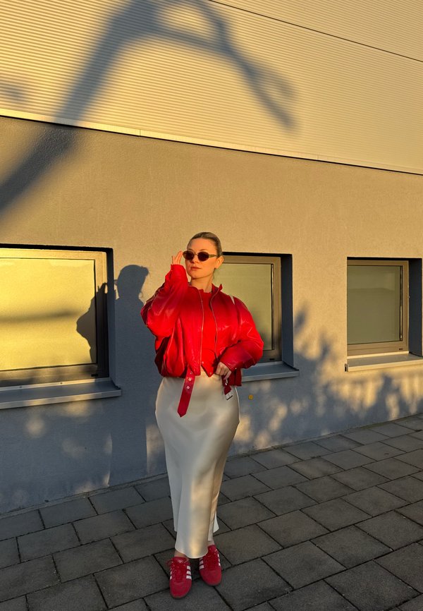 Femme en veste rouge, jupe blanche et baskets rouges ajuste ses lunettes de soleil en se tenant sur un trottoir pavé près d'un bâtiment gris avec des fenêtres.