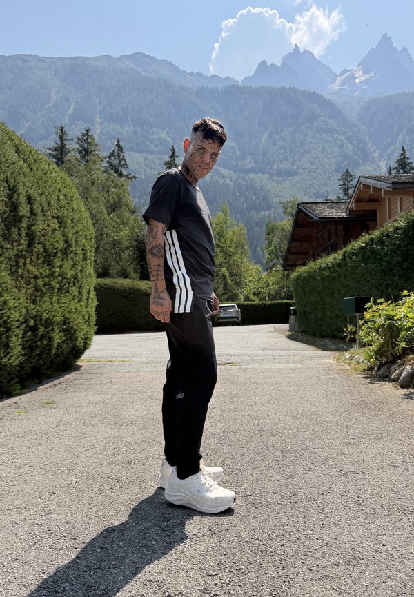 Junger Mann mit Tattoos, der schwarze Sportbekleidung und weiße Turnschuhe trägt, steht auf einer gepflasterten Straße in einer bergigen ländlichen Gegend.