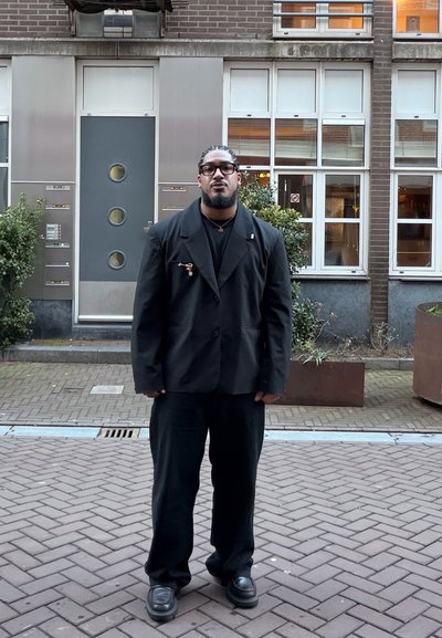 Hombre con cabello trenzado y gafas de pie en una calle pavimentada vistiendo un traje negro, frente a un edificio de ladrillo con ventanas y plantas.