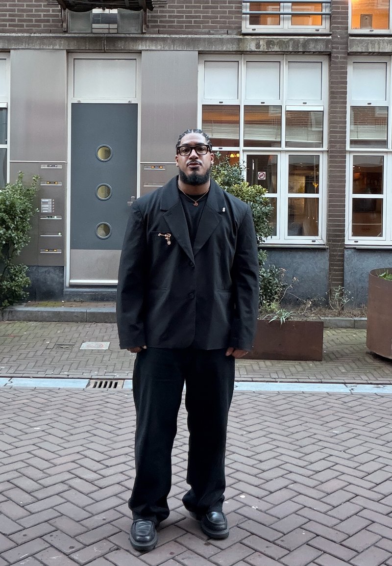 Homme aux cheveux tressés et portant des lunettes, debout dans une rue pavée, vêtu d'un costume noir, devant un bâtiment en briques avec des fenêtres et des plantes.