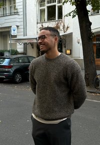 Καφέ, υφαντή πλεκτή μπλούζα πάνω από λευκό πουκάμισο, συνδυασμένη με γκρι παντελόνι. Το άτομο στέκεται έξω, περιτριγυρισμένο από αστικό τοπίο.