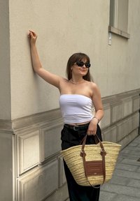 Πλεκτή τσάντα από άχυρο με καφέ δερμάτινες λεπτομέρειες, ορθογώνια βάση και διπλές λαβές. Το μοντέλο φοράει λευκό τοπ και μαύρα παντελόνια.