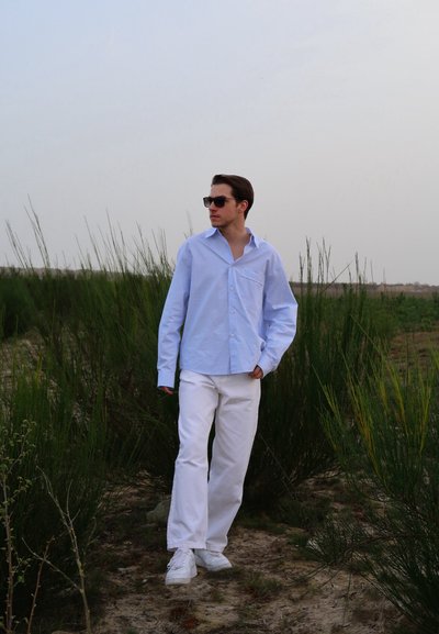 Camisa azul claro de botones y mangas largas, con un bolsillo en el pecho, combinada con pantalones blancos y zapatillas blancas, contra un fondo verde y cubierto de hierba.