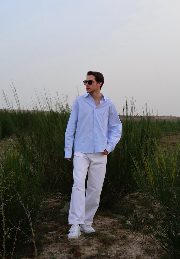 Camisa azul clara de botões, com mangas longas e bolso no peito, combinada com calças brancas e ténis brancos, em fundo de relva verde.