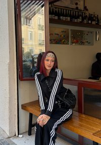 Sort atletisk træningsdragt med hvide striber, siddende på en træbænk, sølvtilbehør synligt, close-up af opmærksomt ansigt i en café.