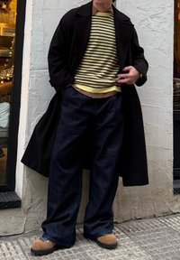 Personne portant un long manteau noir, un pull rayé jaune et noir, un pantalon ample foncé et des chaussures brunes texturées, debout contre un mur clair.