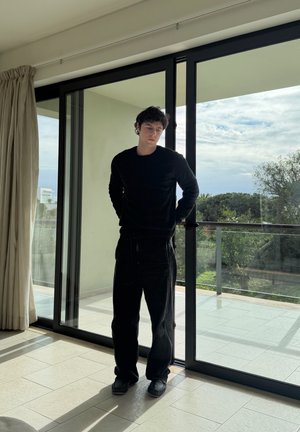 Joven con ropa negra de pie en el interior junto a una puerta corredera de cristal que conduce a un balcón con árboles y cielo nublado afuera.