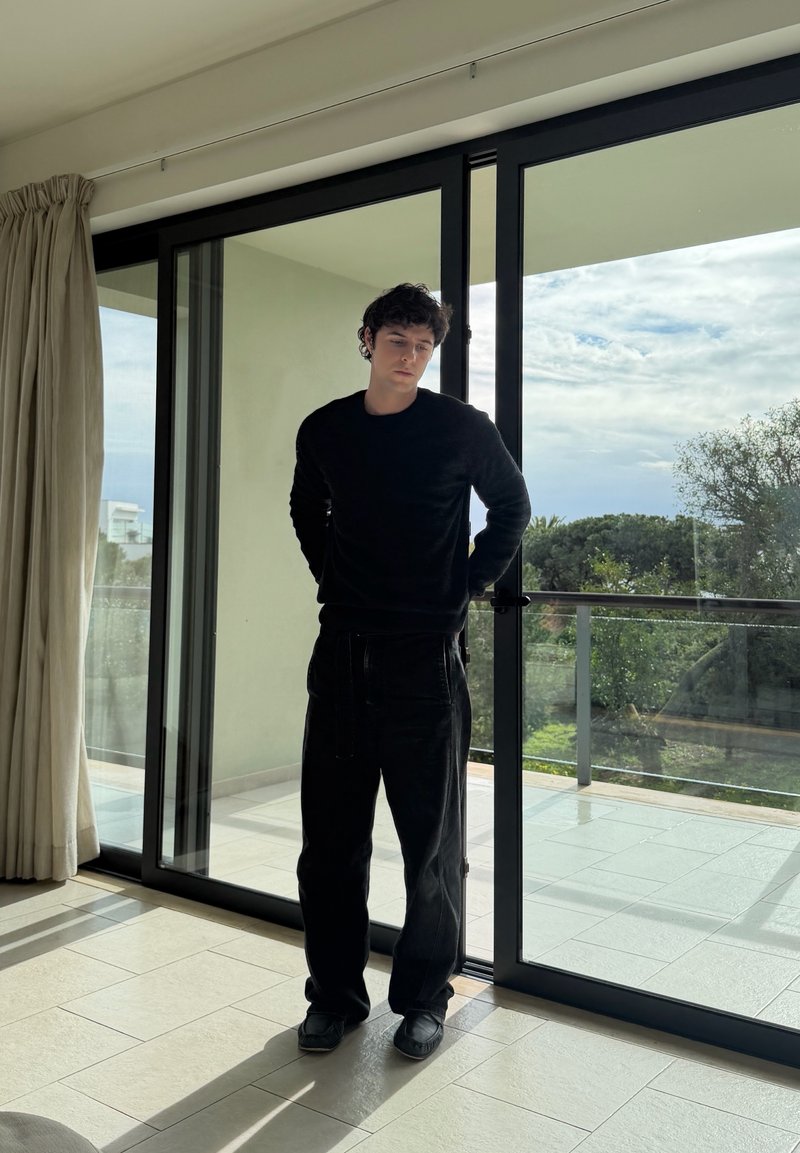 Ung mann i svarte klær står inne ved glass skyvedør som fører til balkong med trær og overskyet himmel utenfor.
