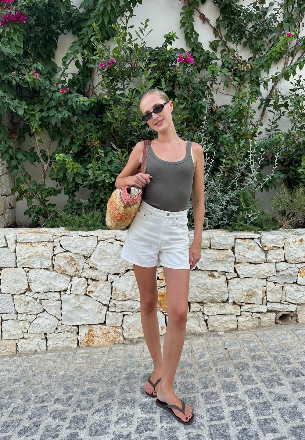 Γκρι φανελάκι, λευκά σορτς και καφέ σανδάλια. Η γυναίκα κρατά μια πολύχρωμη υφασμάτινη τσάντα. Στο φόντο υπάρχει πέτρινος τοίχος και πράσινη βλάστηση.