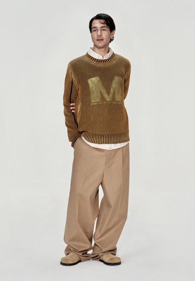 Suéter de punto marrón con ribetes y un gran gráfico amarillo de "M", combinado con pantalones holgados color arena y zapatos slip-on beige. Fondo claro.