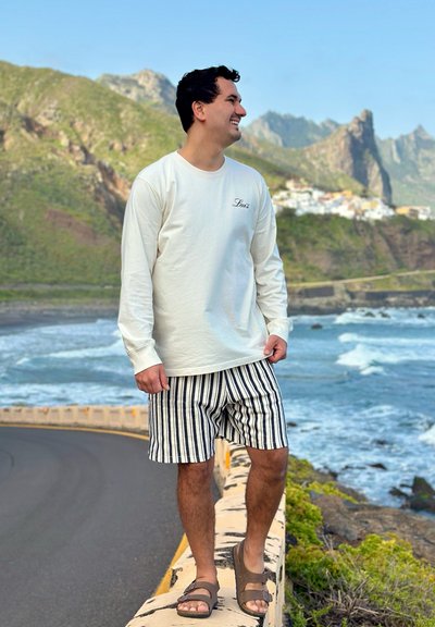 Hombre con camisa blanca de manga larga y pantalones cortos a rayas de pie sobre una barrera al borde de la carretera junto al océano, con montañas y un pueblo costero en el fondo.