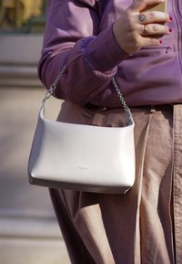 Borsa a tracolla in pelle grigia chiara con catena, caratterizzata da un design semplice e un logo dorato. La borsa ha una texture liscia e una forma strutturata.