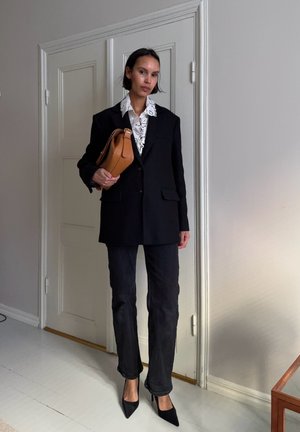 Mujer con blazer negro de gran tamaño, blusa blanca de encaje, pantalones negros, tacones negros, sosteniendo un bolso marrón en una habitación con puerta blanca cerrada.