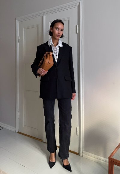 Mujer con blazer negro de gran tamaño, blusa blanca de encaje, pantalones negros, tacones negros, sosteniendo un bolso marrón en una habitación con puerta blanca cerrada.