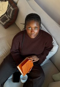 Femme aux cheveux tressés portant un pull marron assise sur un canapé gris, tenant un livre orange ouvert intitulé "Manifest", avec une tasse et un téléphone à proximité.