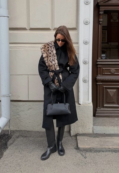 Mujer con abrigo negro, bufanda de piel estampado de leopardo, guantes y botas negras, sosteniendo un pequeño bolso negro, de pie junto a una pared beige y una puerta marrón.